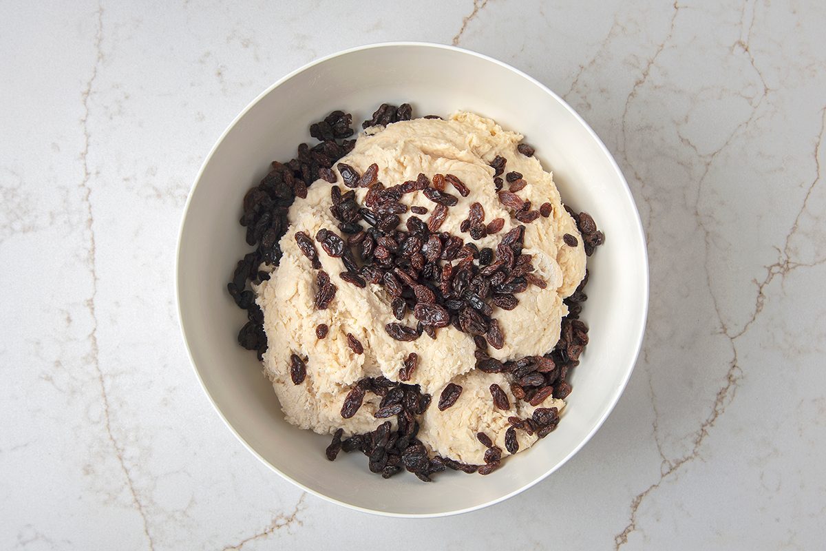 A bowl of light brown dough topped with raisins on a marble countertop.