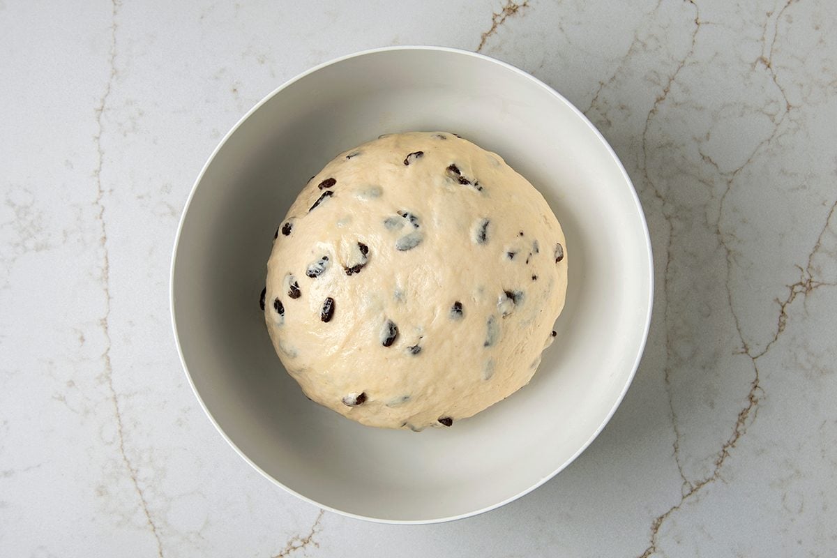 A round ball of bread dough with chocolate chips is set inside a white bowl. The bowl is placed on a light, marbled countertop.