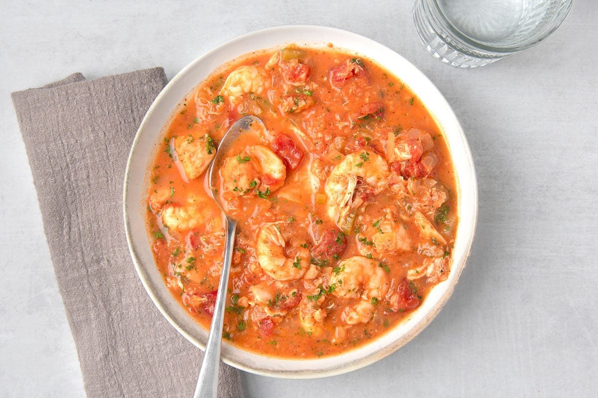 overhead shot of a bowl of seafood stew; the stew is a rich red color and contains large chunks of shrimp, fish, and vegetables; a spoon is in the bowl and a glass of water is next to the bowl; the bowl is on a light gray surface with a gray napkin to the left of the bowl