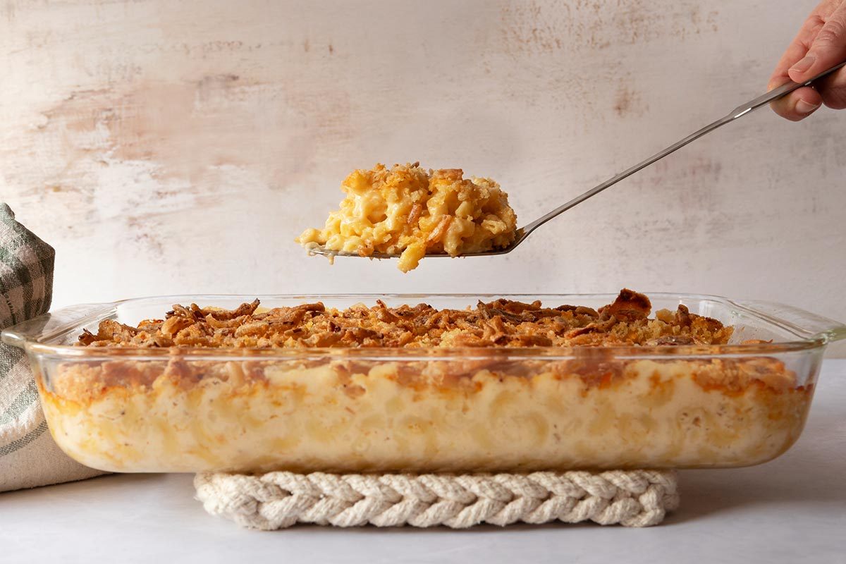 A large glass baking dish full of cooked mac and cheese rests on a braided potholder on a countertop, with a stone background and a hand holding a spatula full of the mac and cheese above the dish