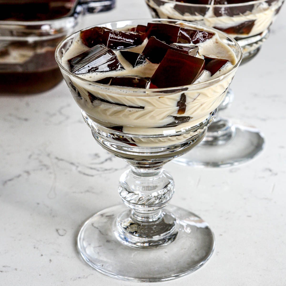 A glass dessert cup filled with cubes of coffee jelly and creamy white sauce, set on a marble surface. The dessert appears elegant and is served in a decorative glass, with another similar serving visible in the background.