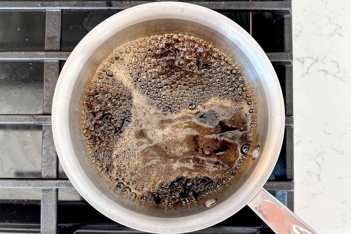 A small saucepan on a stove with liquid gently bubbling inside, appearing to be boiling coffee or a dark sauce. The stove features a black burner grate. The countertop is light-colored and speckled.