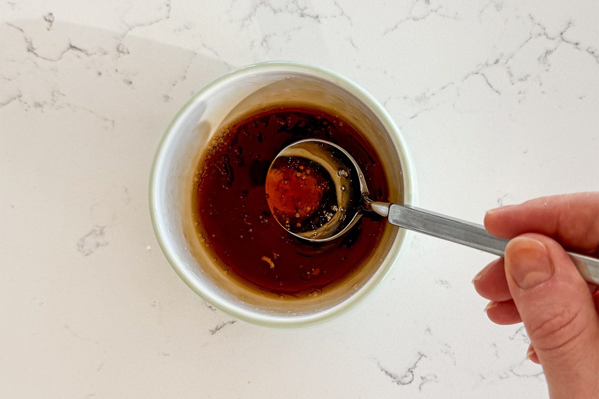 A hand holds a spoon over a bowl containing brown liquid, possibly a sauce or dressing, with visible herbs and spices. The bowl is placed on a white marble countertop.