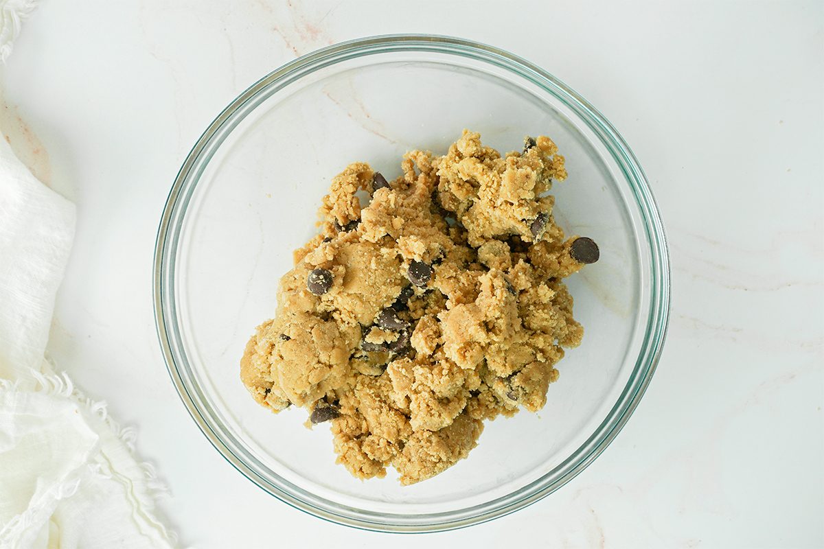A glass bowl filled with chocolate chip cookie dough on a light-colored surface. A white cloth is partially visible on the left side.