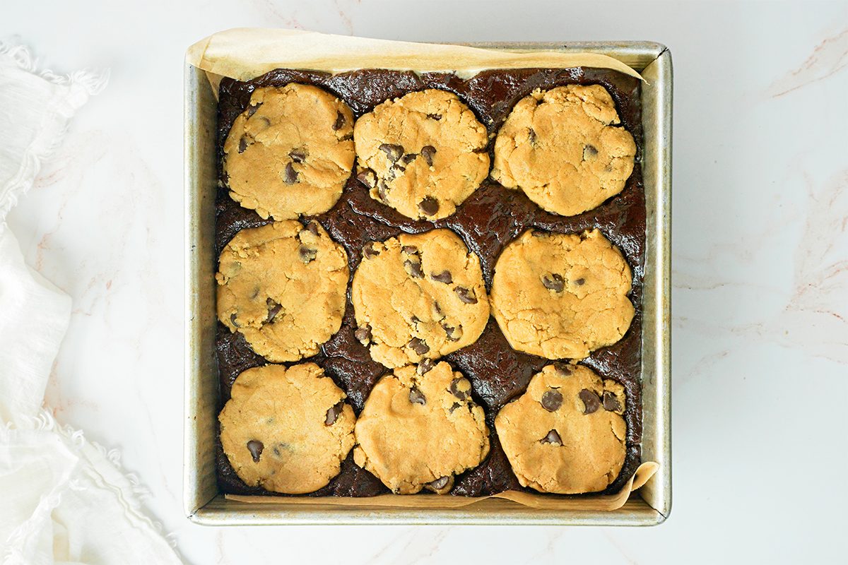 A square baking pan filled with layered dessert bars featuring a dark brownie base topped with nine round cookie dough portions, embedded with chocolate chips. The pan is lined with parchment paper on a light marble surface.