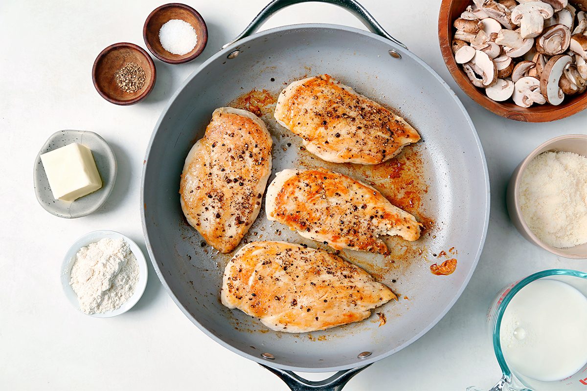 A pan with four cooked chicken breasts seasoned with pepper. Surrounding the pan are ingredients including bowls of mushrooms, flour, butter, salt, pepper, and a measuring cup with milk, arranged on a light surface.