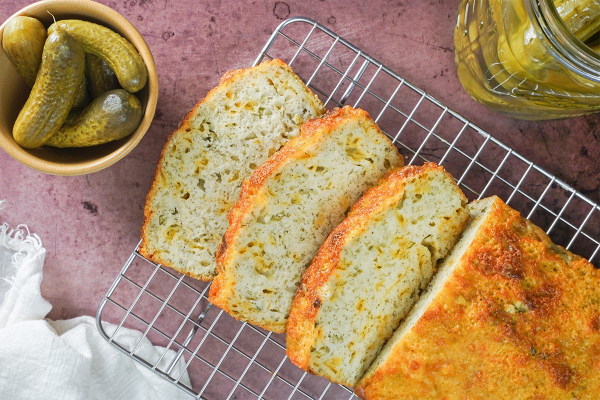Sliced dill pickle bread on a wire rack with a bowl of pickles and a jar of pickles nearby, all on a pink surface.