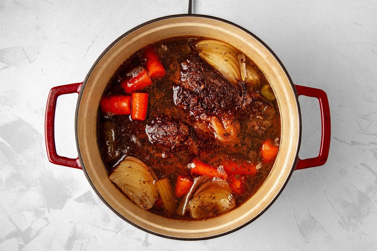 overhead shot of a Dutch oven pot roast cooking on the stovetop; a Dutch oven pot roast is a slow-cooked dish made with beef, vegetables, and broth; the pot is filled with a brown liquid that is likely broth or gravy, and there are chunks of meat, carrots, and onions in the liquid