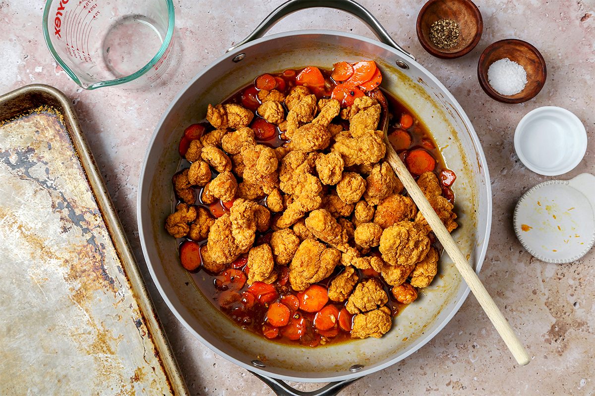 A pan filled with cooked pieces of seasoned chicken and sliced carrots. A wooden spoon rests on the pan. Surrounding the pan are small bowls with seasonings, a measuring cup, and a baking sheet on a textured surface.