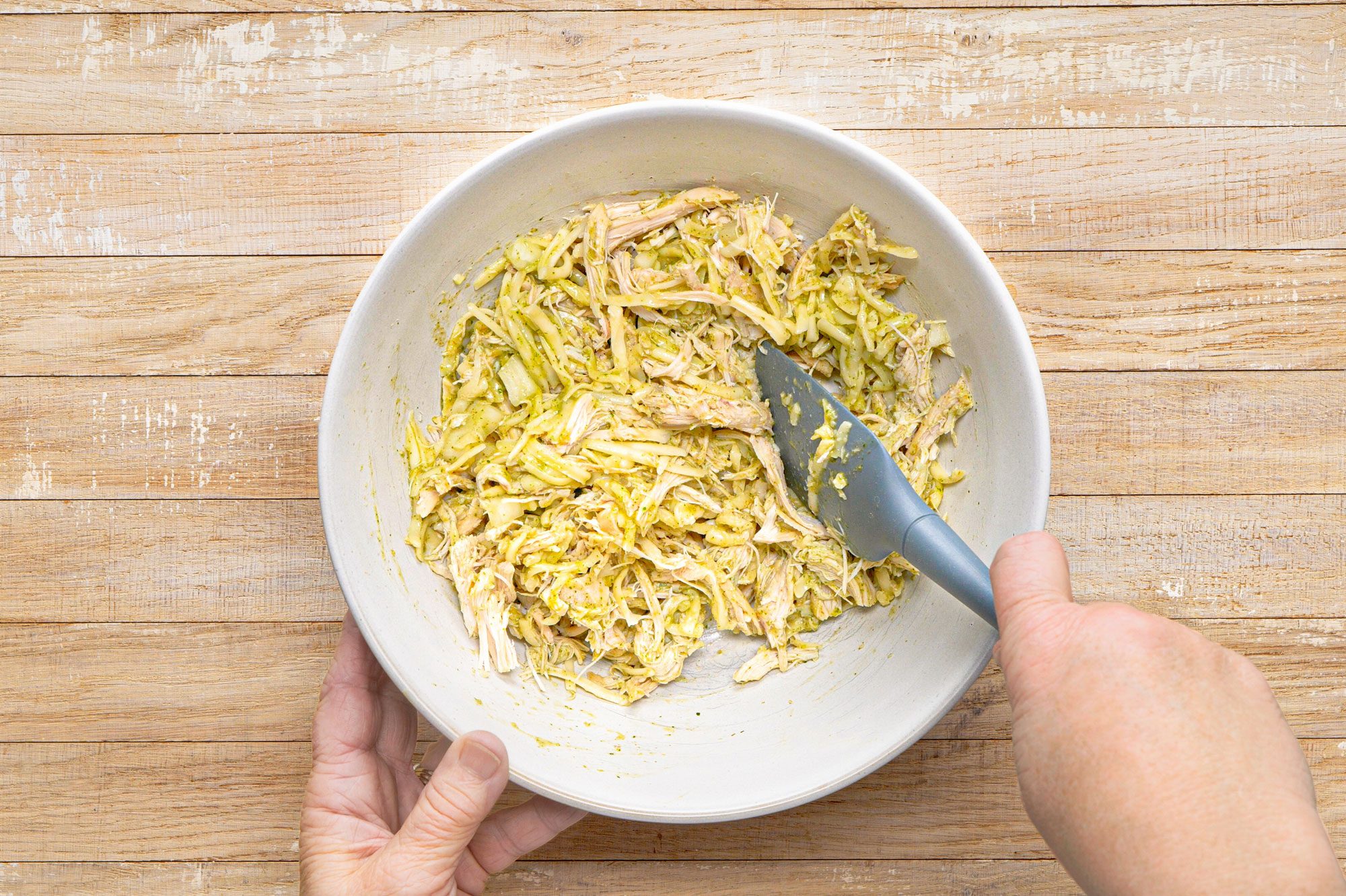 overhead shot of a white bowl filled with shredded chicken and a green sauce is being stirred with a blue spatula; a hand is holding the bowl from below and another hand is holding the spatula; the bowl is placed on a wooden surface