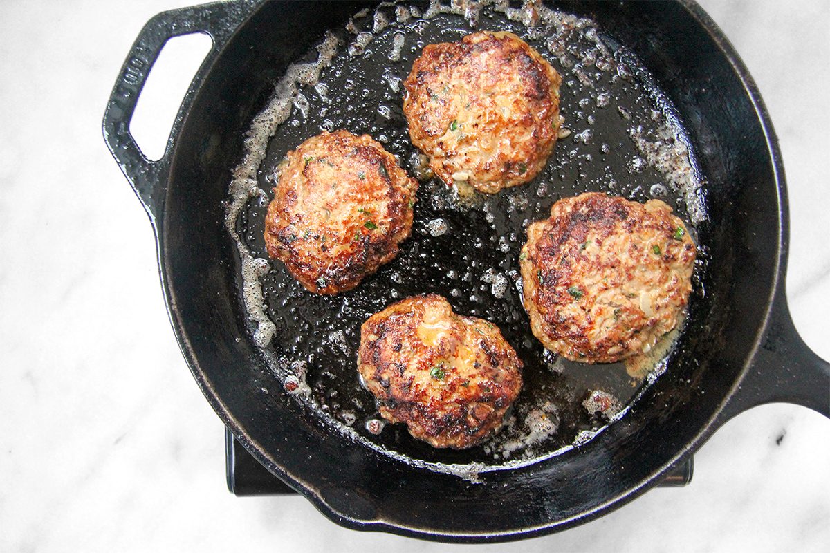 Four golden-brown patties sizzling in a black cast-iron skillet on a marble surface.