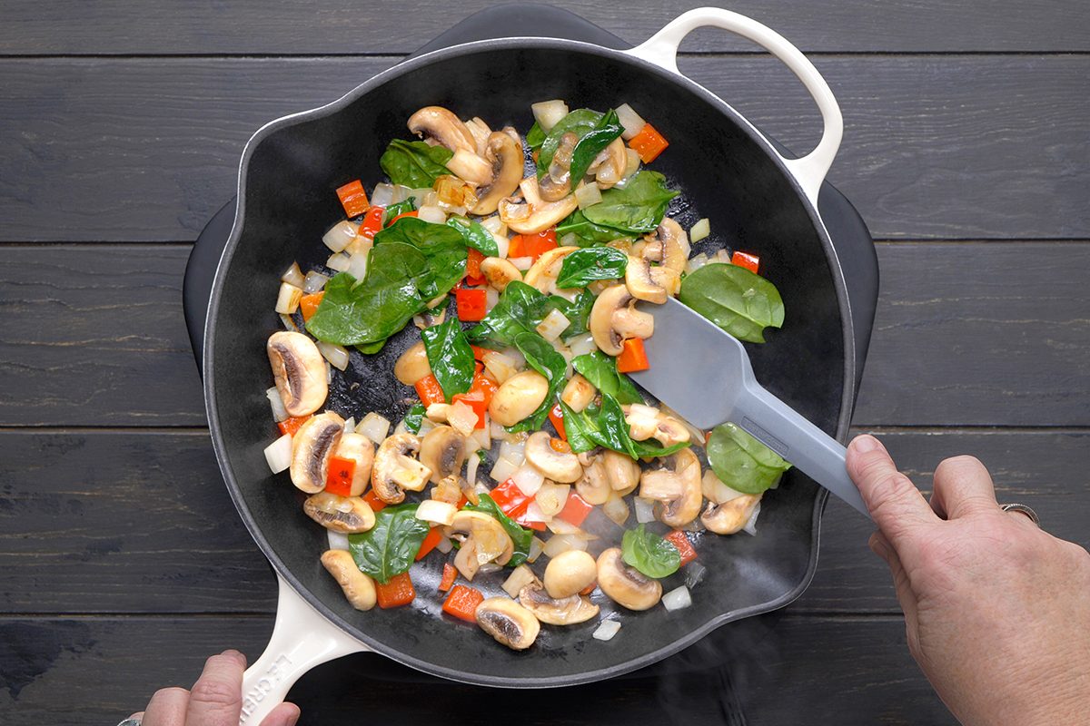 A person sautés mushrooms, spinach, red bell peppers, and onions in a pan using a gray spatula. The pan is on a black stovetop with steam rising from the vibrant vegetables.