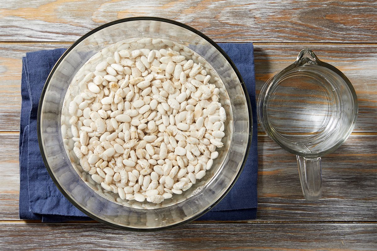 A glass bowl filled with white beans soaking in water sits on a wooden surface with a blue cloth underneath. Next to it, there is an empty glass measuring cup.