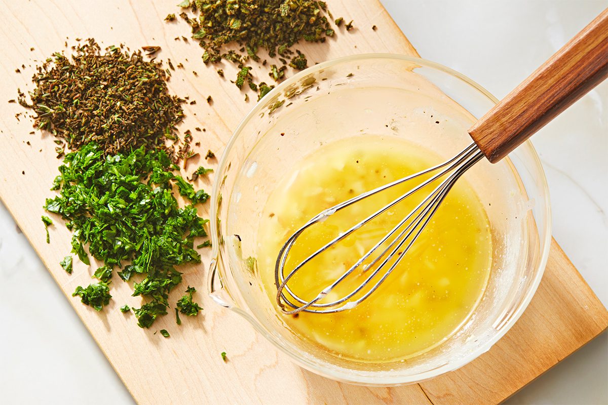 A glass bowl filled with a yellow liquid and a whisk on a wooden board. Surrounding the bowl are small piles of chopped fresh herbs, including parsley and dried herbs. The board is placed on a light-colored surface.
