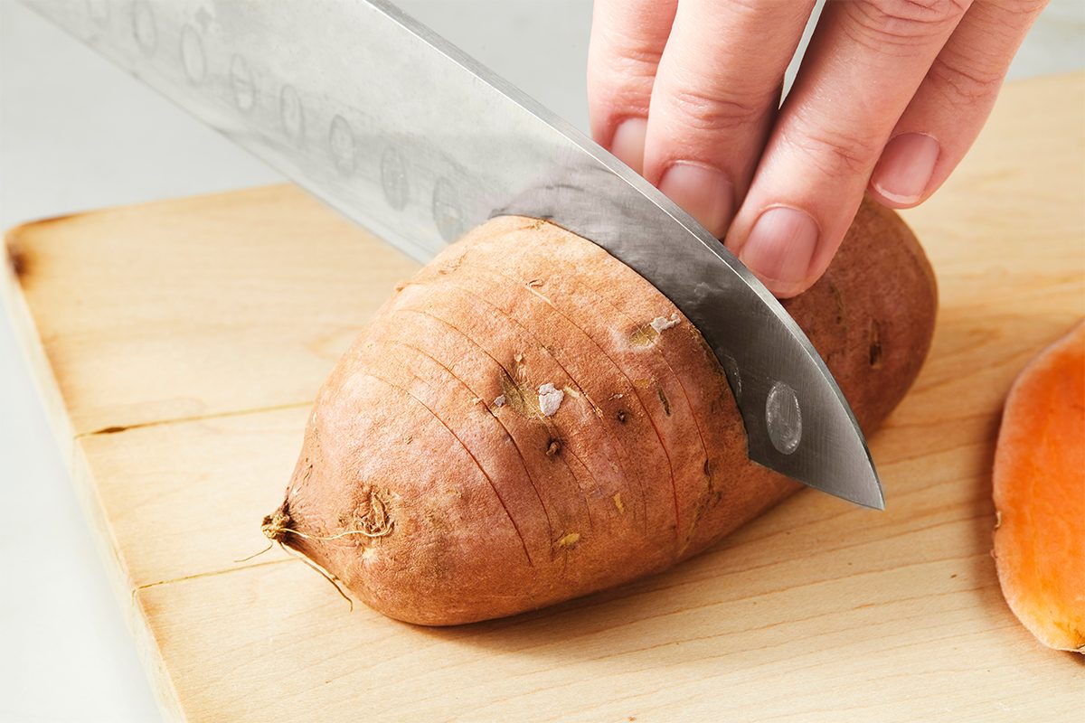 A hand holding a knife is cutting a sweet potato on a wooden cutting board. The sweet potato is partially sliced, and some of its orange interior is visible.
