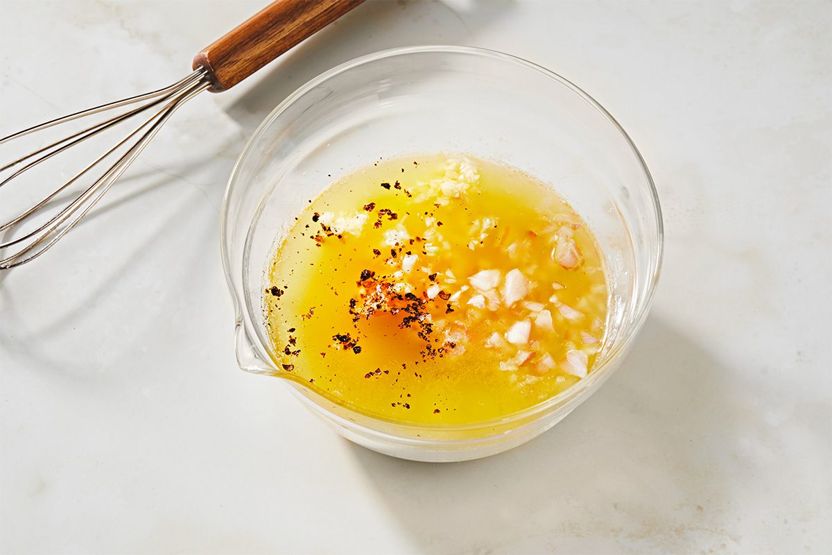 A clear glass bowl contains a yellow mixture of oil, finely chopped garlic, and black pepper, set on a light-colored marble surface. A whisk with a wooden handle lies beside the bowl.