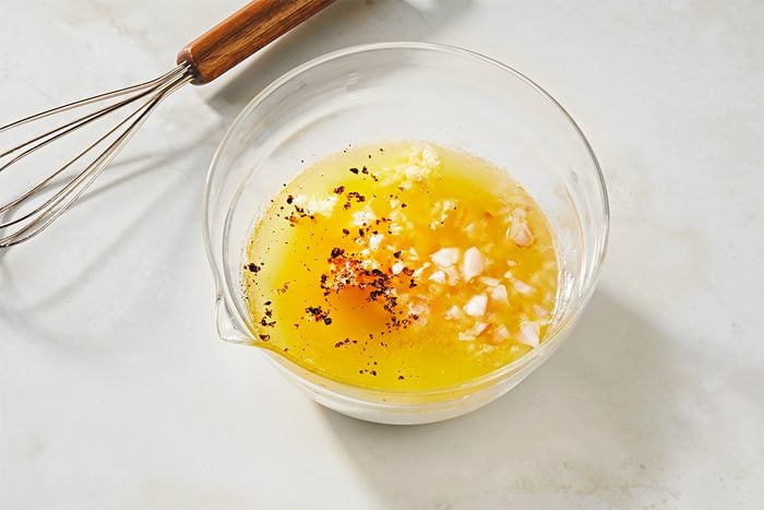 A clear glass bowl contains a yellow mixture of oil, finely chopped garlic, and black pepper, set on a light-colored marble surface. A whisk with a wooden handle lies beside the bowl.