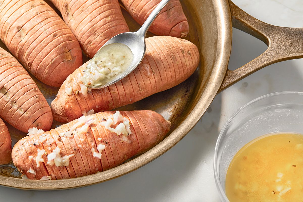 A spoon is applying a garlic butter mixture onto sliced sweet potatoes in a pan. A small bowl with remaining garlic butter is nearby on a marble surface.