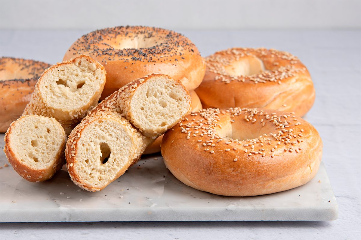 A variety of bagels are displayed on a white marble surface. Some are topped with sesame seeds, others with poppy seeds, and one is sliced in half to show the texture. The background is a light gray.