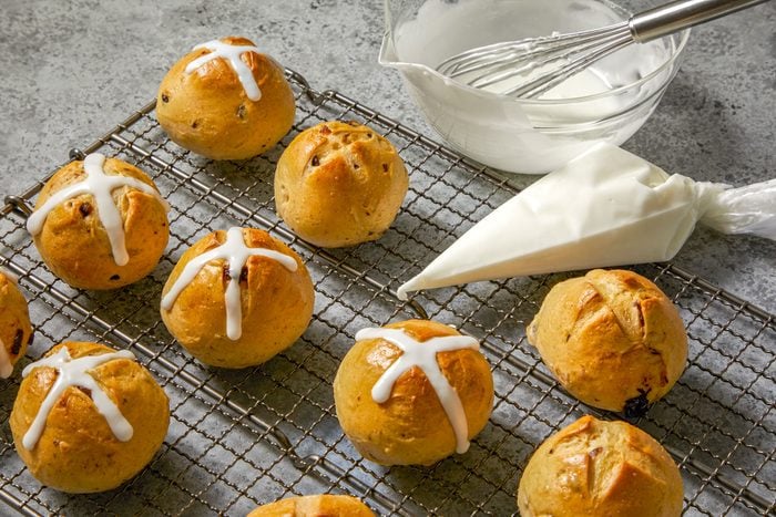 3/4th shot of a plate of Traditional Hot Cross Buns; the buns are arranged in a criss-cross pattern on a wire rack, with a few buns in the foreground and a few in the background; the buns are surrounded by a bowl of white icing, a piping bag of white icing, and a whisk