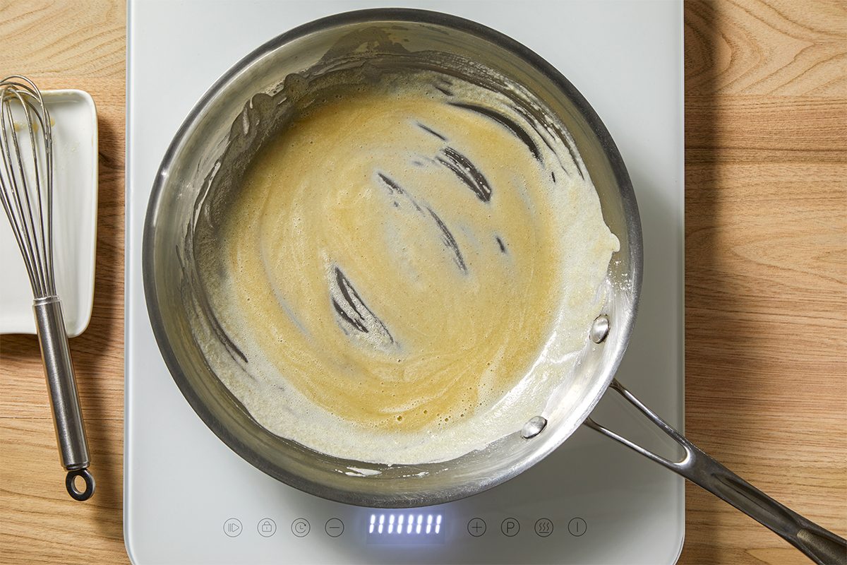 A stainless steel pan on a white induction cooktop contains a light brown roux being stirred. A metal whisk is placed nearby on the wooden countertop.