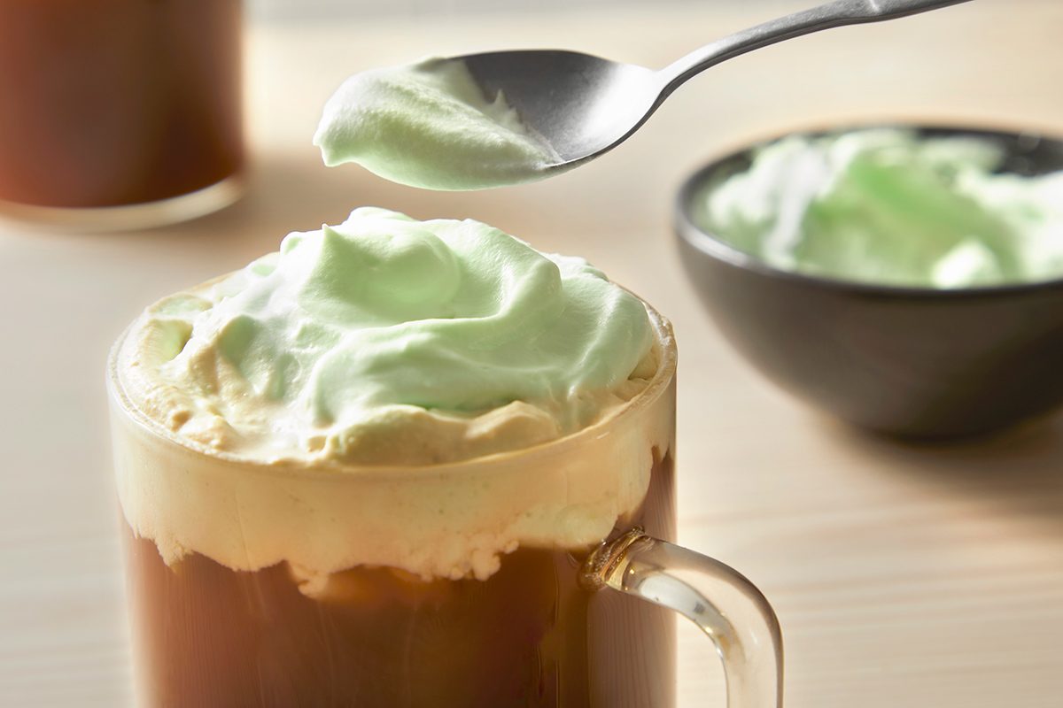 A glass mug filled with coffee topped with frothy green whipped cream. A spoon is lifting a portion of the cream above the mug. A bowl containing more green whipped cream is in the background on a wooden surface.