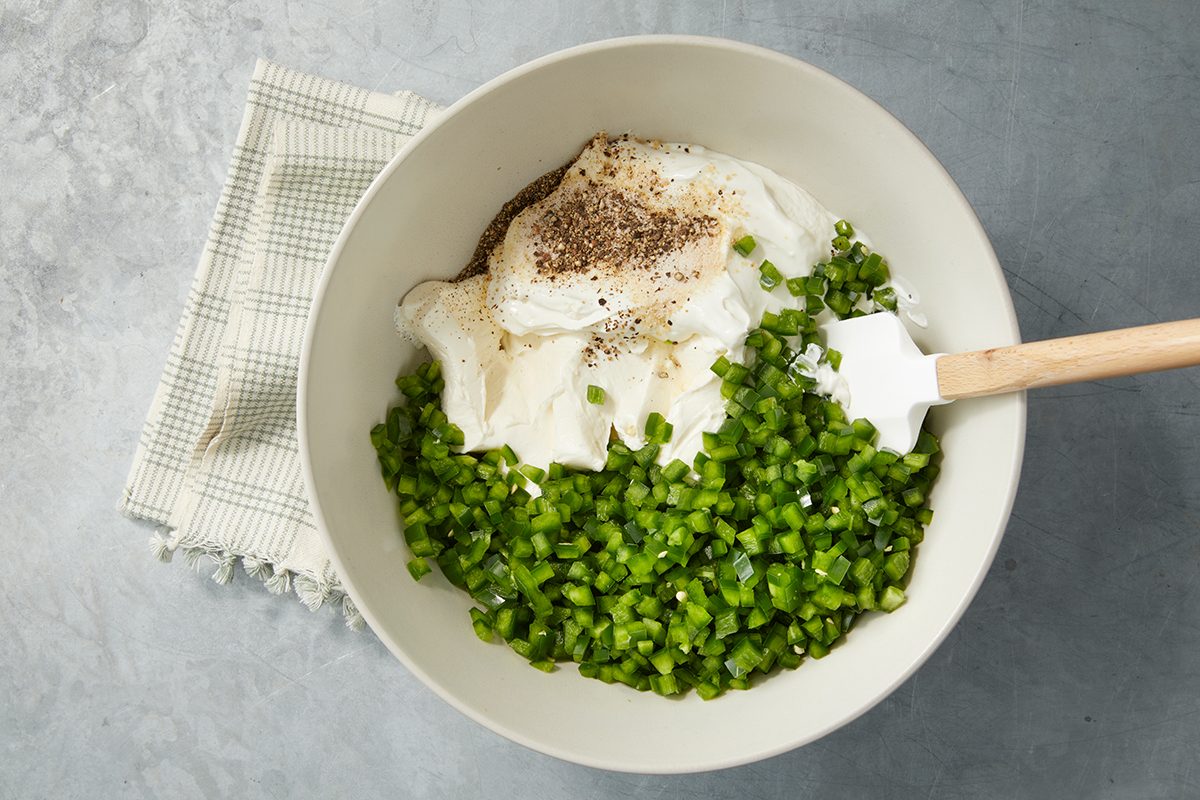 Bowl containing chopped green peppers, mayonnaise, and black pepper with a spatula, placed on a checkered cloth on a gray surface.