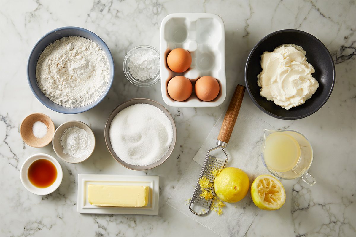 Overhead view of ingredients required to make lemon ricotta cake on white marble countertop.