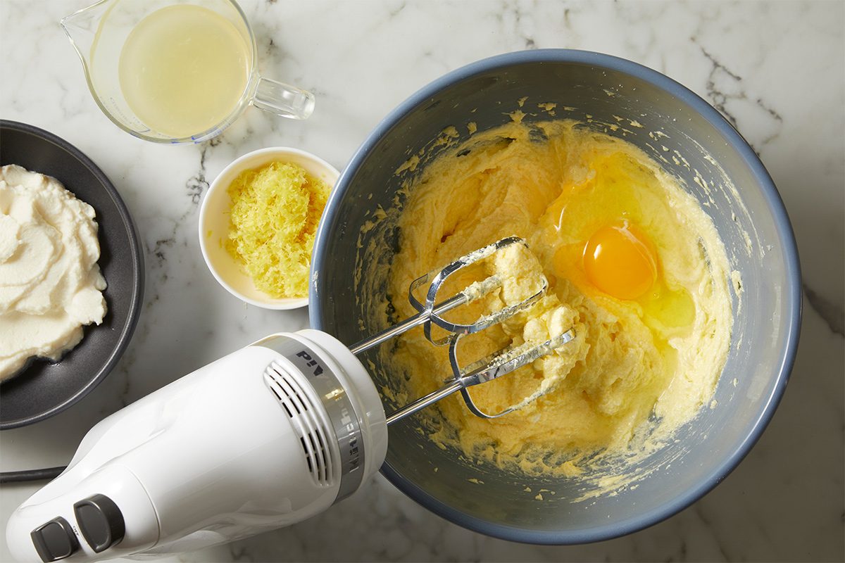 Overhead view of a bowl with an egg being whisked into softened butter on a white marble countertop. Next to it is are small bowls containing ricotta, lemon juice and lemon zest.