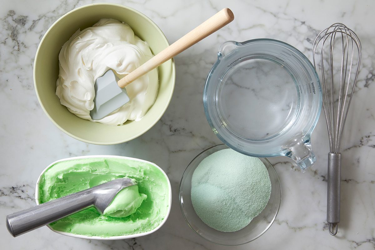 Overhead view of ingredients required to make lime gelatin salad on a white marble countertop with a whisk kept beside it.