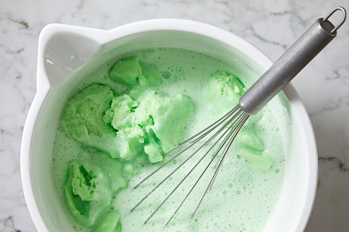 Overhead view of a large white bowl with a whisk with the lime sherbet being whisked into the lime gelatin mixture on a white marble countertop.