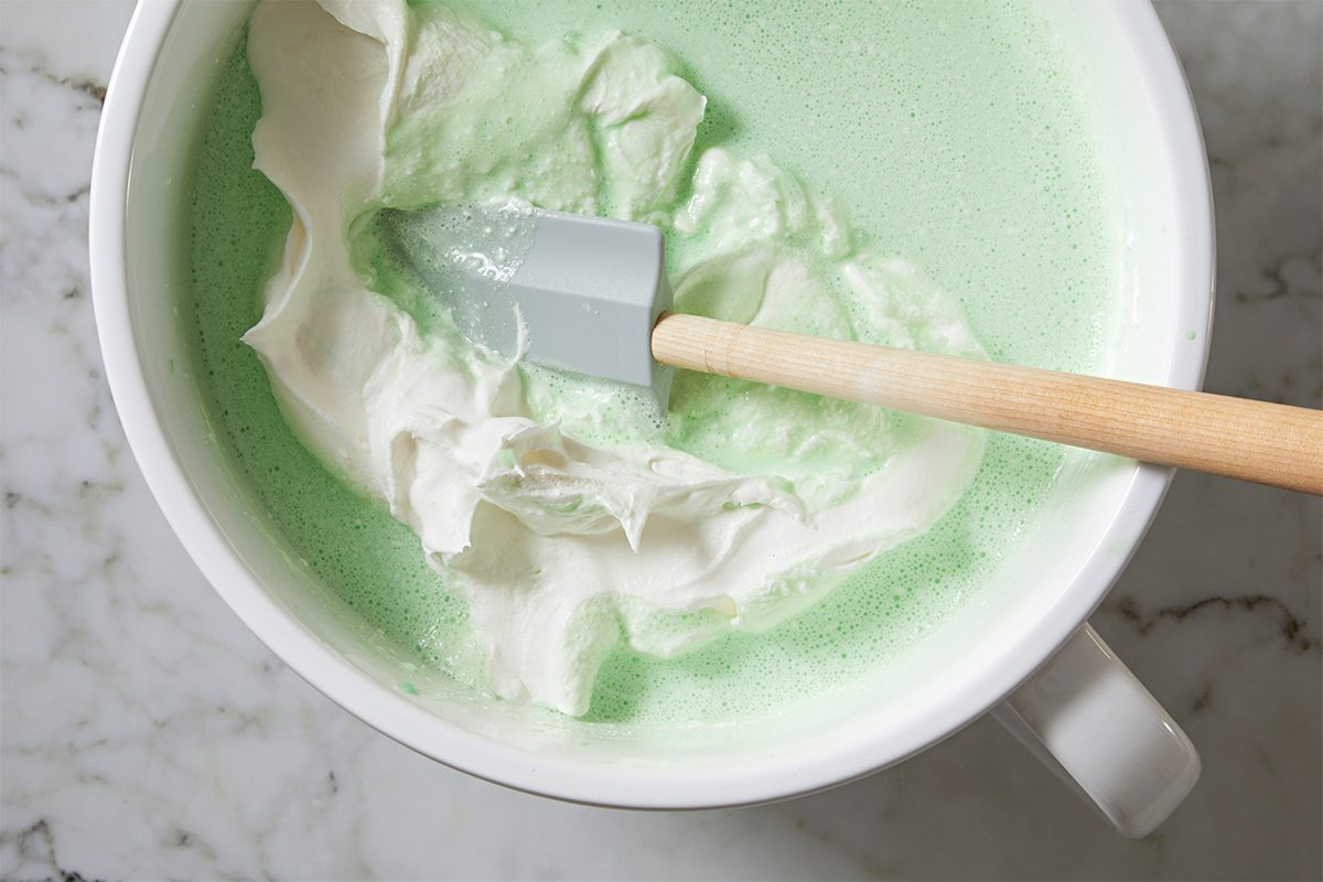 Overhead view of a large white bowl with a wooden spatula with the whipped topping being whisked into the lime gelatin and sherbet mixture on a white marble countertop.