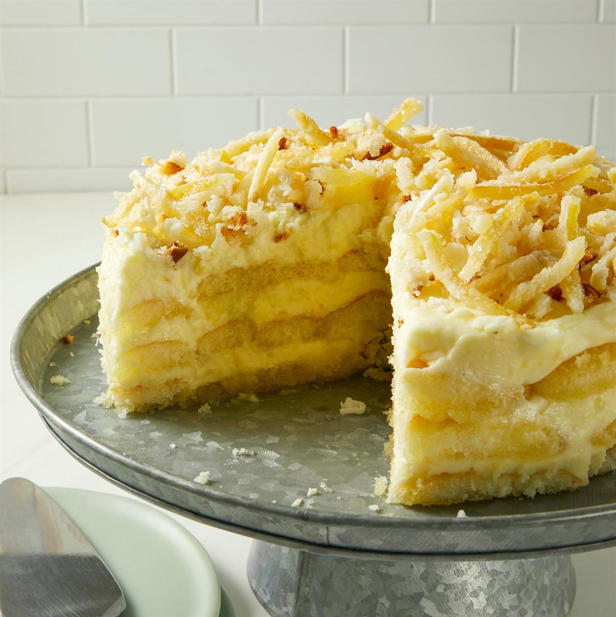 A layered cake with white frosting is displayed on a metal plate. The cake is topped with shredded coconut and toasted nuts. One slice has been removed, revealing the layers. Plates and a server are visible beside the cake.