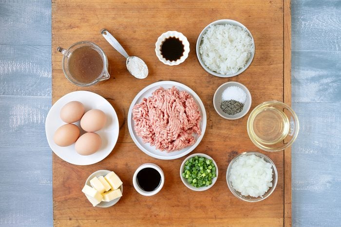 Ingredients neatly arranged on a wooden surface, including ground meat, eggs, chopped green peppers, diced onions, butter, sauces, seasoning, and liquids in small bowls and plates.