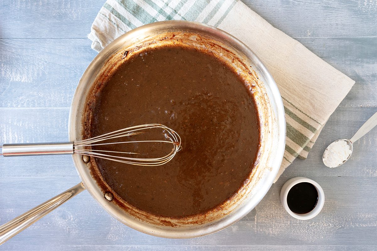 A saucepan filled with dark brown sauce being whisked. It is placed on a light blue surface with a striped cloth nearby. A small cup of dark liquid and a spoon with a white substance are also visible beside the pan.