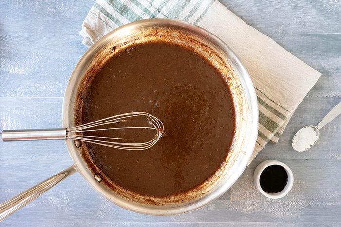 A saucepan filled with dark brown sauce being whisked. It is placed on a light blue surface with a striped cloth nearby. A small cup of dark liquid and a spoon with a white substance are also visible beside the pan.