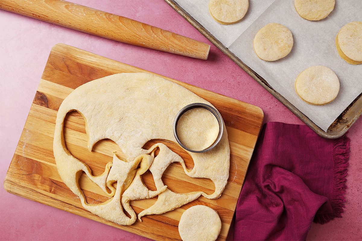 A wooden board with rolled-out dough and a cookie cutter is shown. Circular dough cut-outs are visible, with one placed on a nearby baking sheet lined with parchment paper. A rolling pin and red cloth are also on the pink surface.
