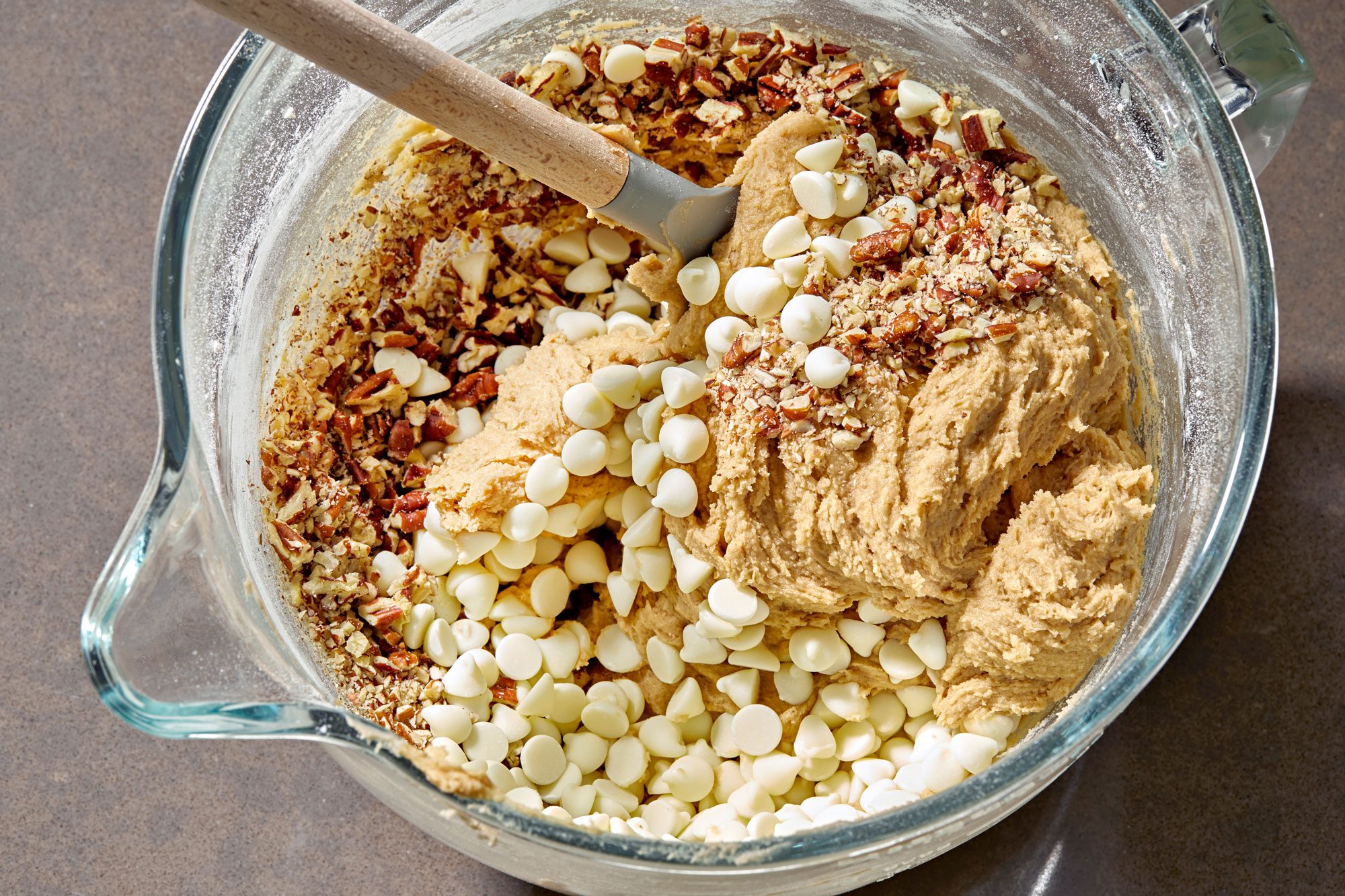 A glass bowl containing cookie dough; a wooden spatula is still resting on top of the cookie dough; The dough is mixed with chopped pecans and white chocolate chips; the sides of the bowl are coated with a thin layer of flour