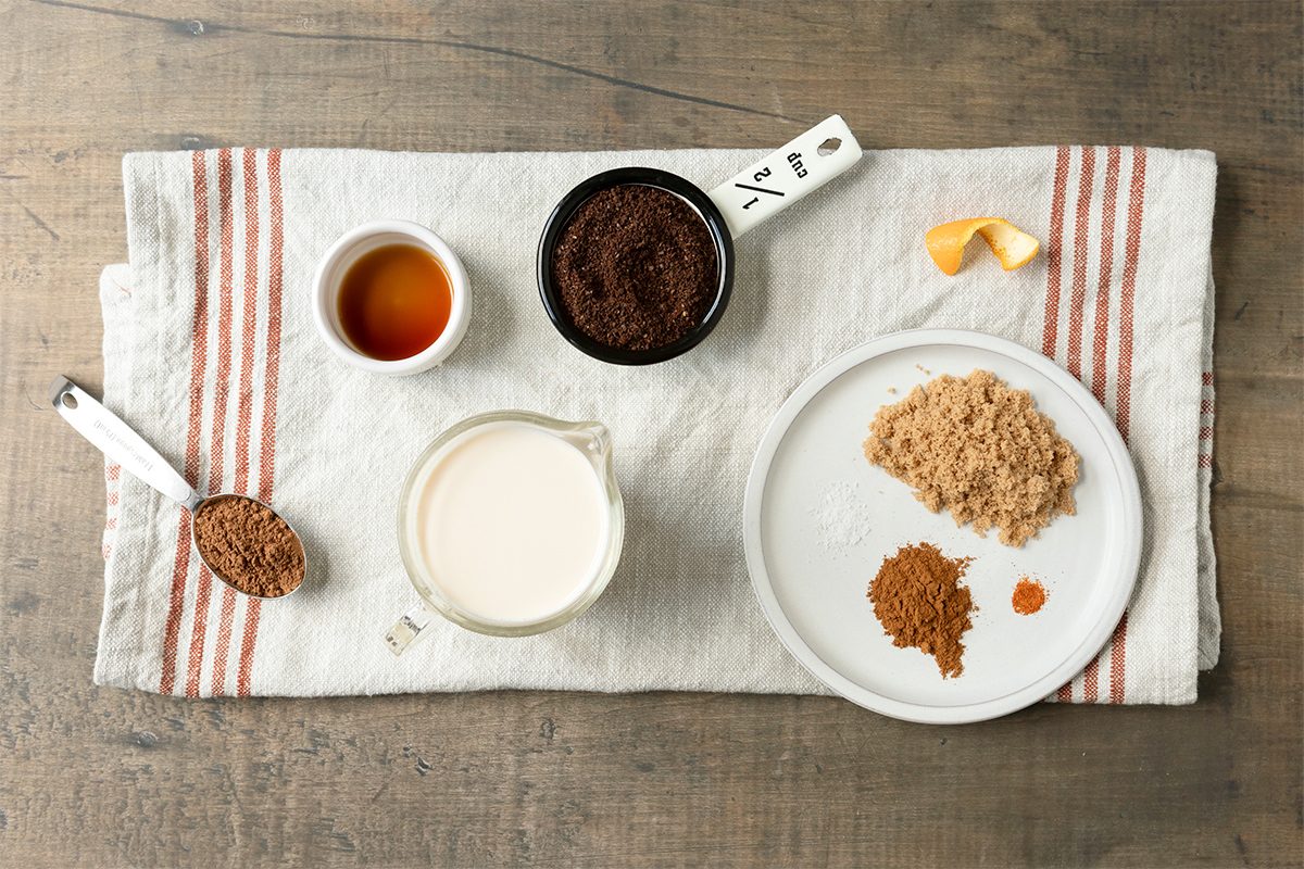 A flat lay of baking ingredients on a striped cloth. Includes a spoon with brown powder, a cup of milk, bowls with dark powder and honey, a plate with brown sugar and spices, and a piece of orange peel.