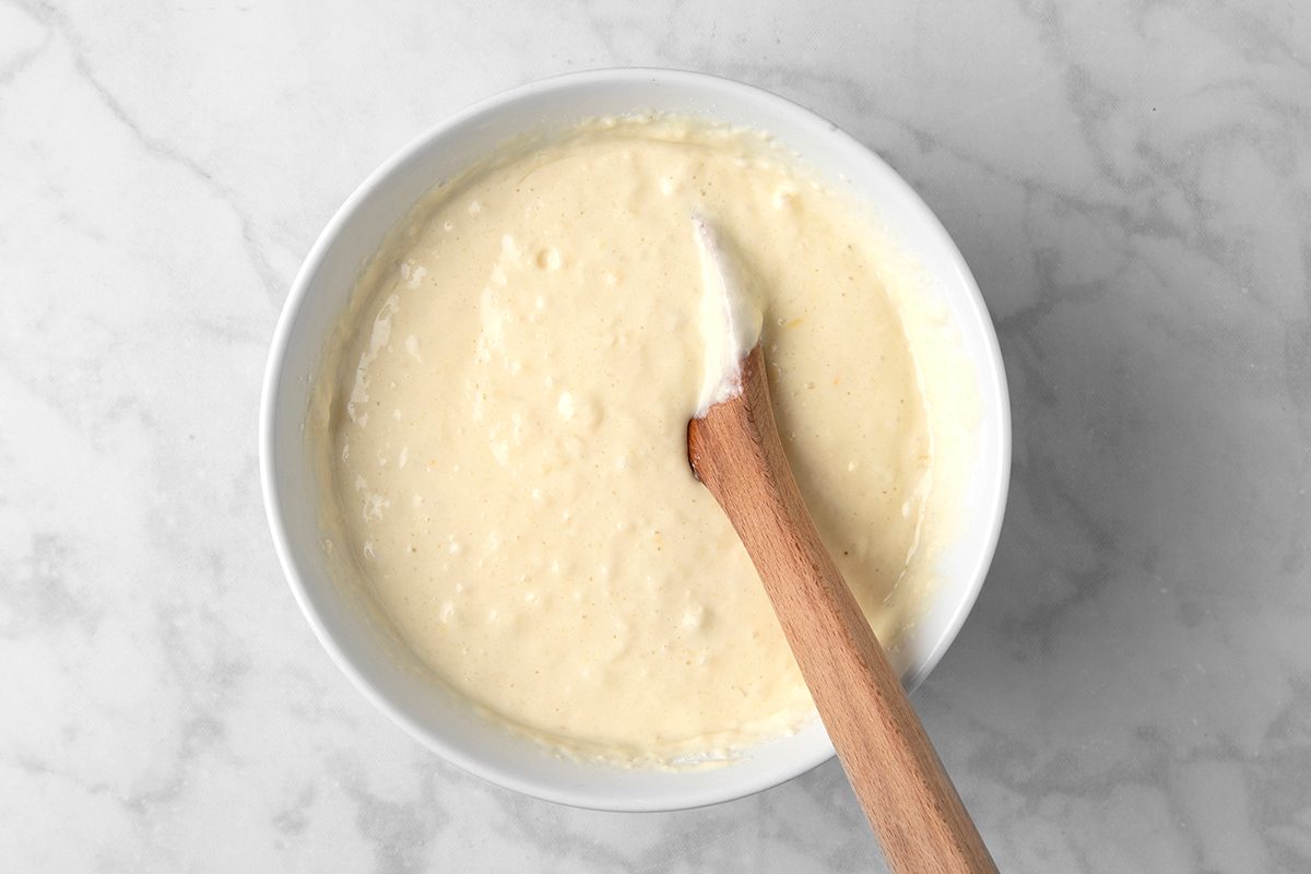A white ceramic bowl filled with creamy, pale yellow batter is placed on a marble countertop. A wooden spoon rests inside the bowl, partially coated with the batter.