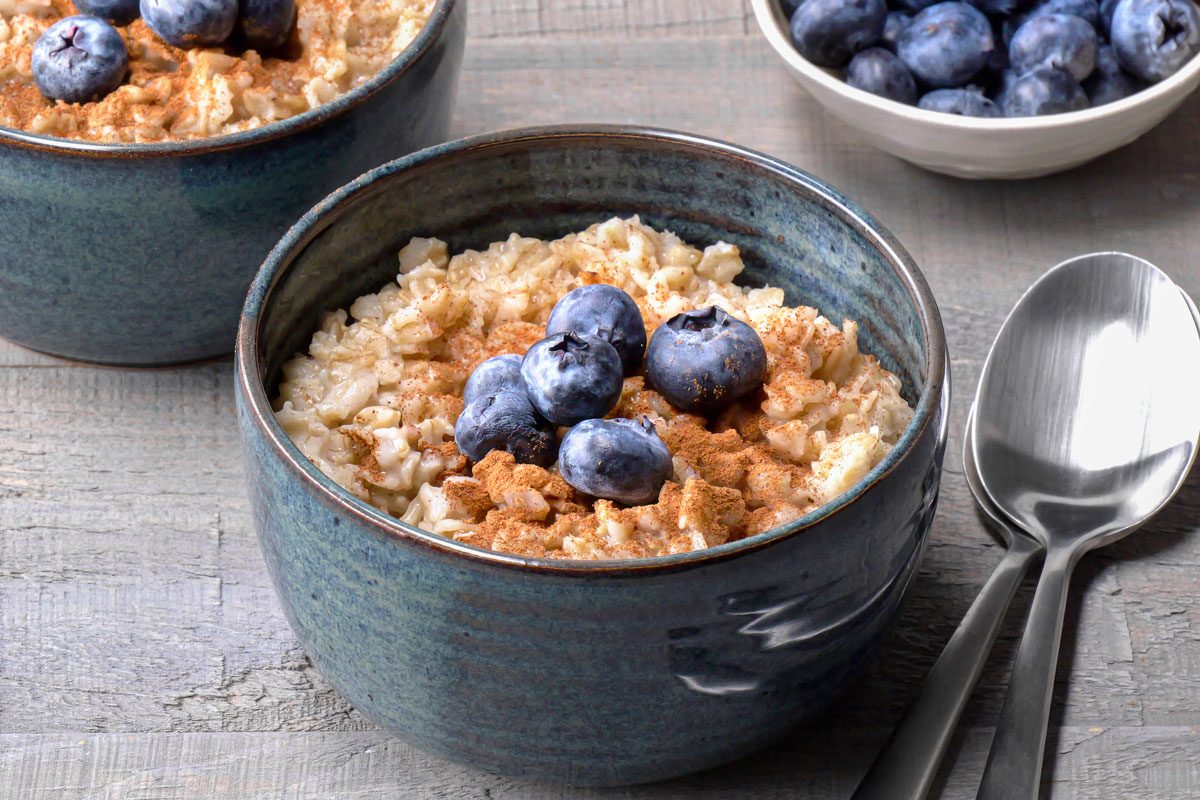 closeup shot of two bowls of oatmeal; one with a spoon and one with a bowl of blueberries, sitting on a wooden table; The oatmeal is topped with blueberries and cinnamon; The bowls are blue and the spoons are silver