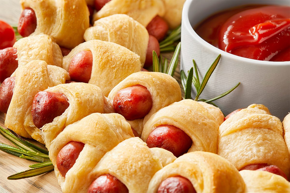 Horizontal AP Push shot of a ring of piggies is arranged on a wooden cutting board, decorated with sprigs of rosemary and cherry tomatoes; a bowl of ketchup is placed in the center of the wreath