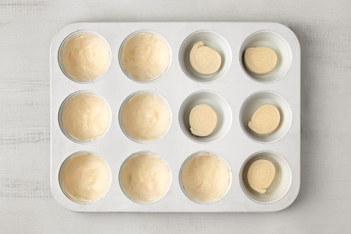overhead shot of a white muffin tin with twelve empty muffin cups; the muffin tin sits on a white wooden surface, the cups are filled with unbaked pie crusts; some of the cups have flat disks of pie crust in them and some of the cups have a hollowed out pie crust ready to be filled with filling