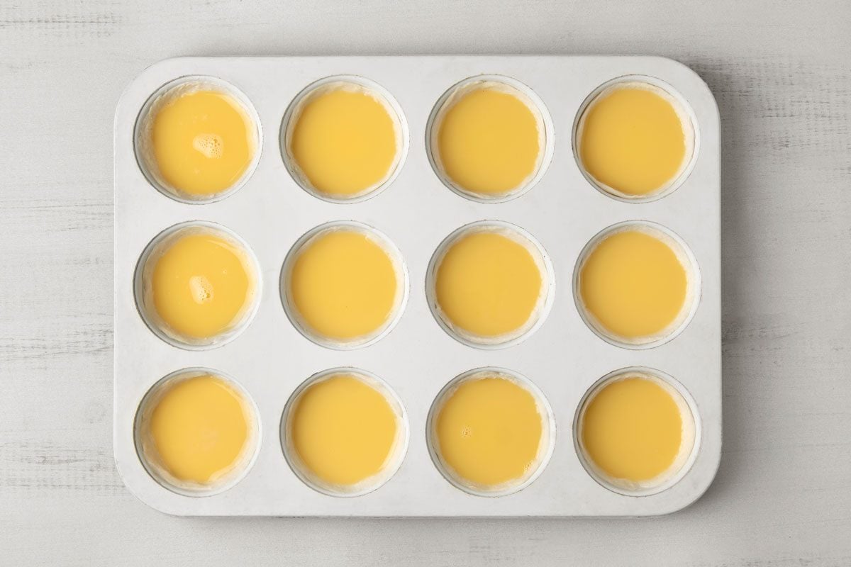 overhead shot of a white, rectangular baking tray with twelve circular depressions filled with a yellow custard-like mixture; the tray is sitting on a white countertop with subtle grey streaks
