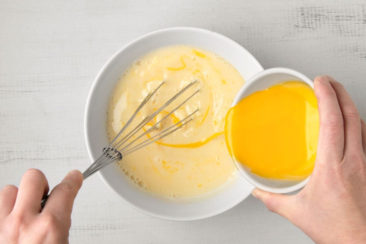 overhead shot of a hand is pouring the contents of a small white bowl into a larger white bowl containing a mixture of milk and egg yolks; the larger bowl also has a whisk in it, and the hand holding the small bowl