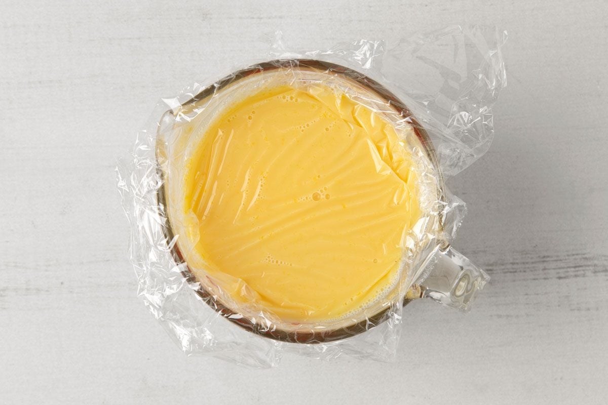 overhead shot of a glass bowl containing a yellow liquid covered with plastic wrap ; the bowl is resting on a white, wooden surface