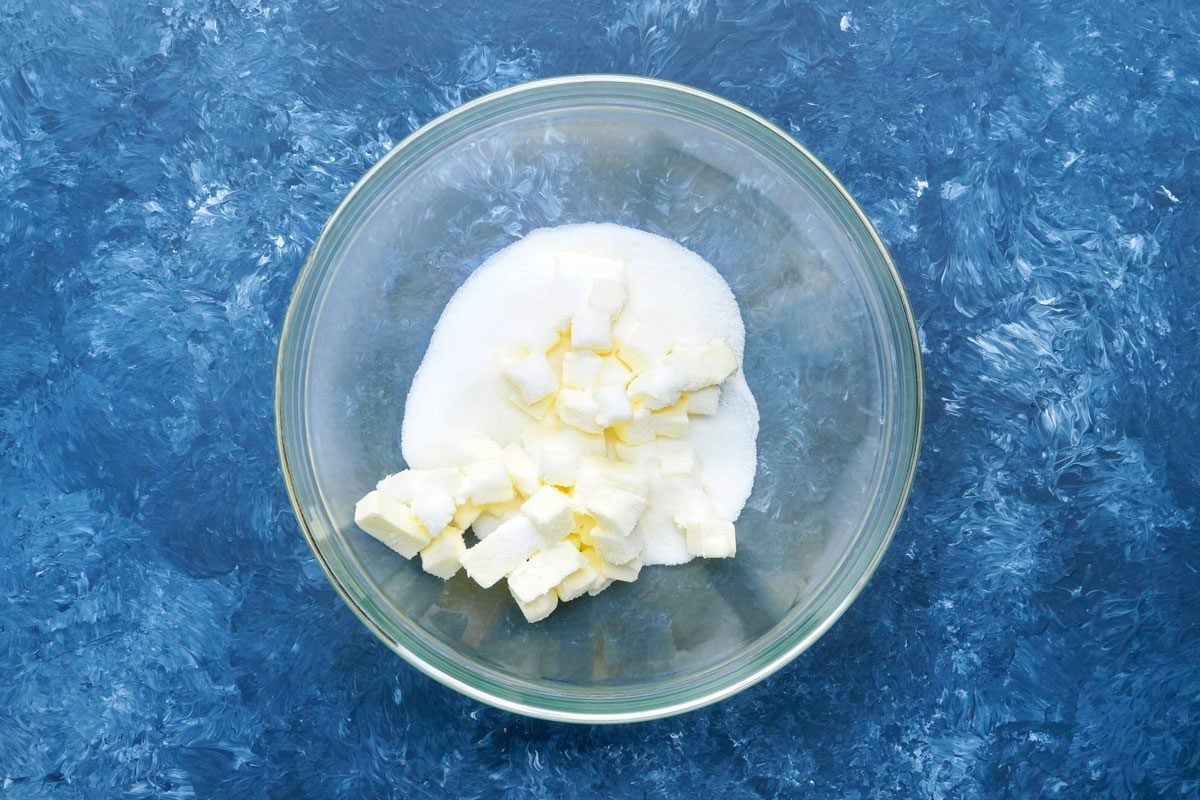 overhead shot of a glass bowl containing cubed butter and granulated sugar; the bowl is sitting on a blue surface with white swirl pattern