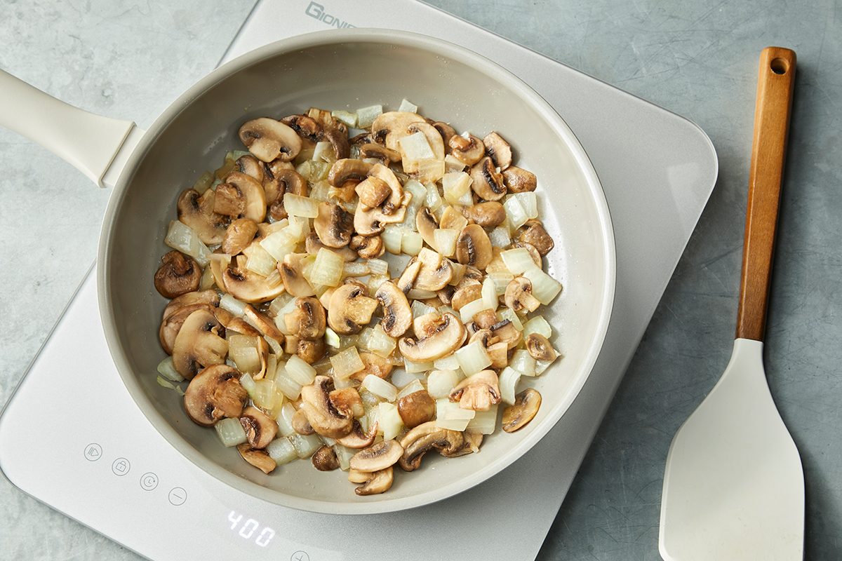 A white frying pan on an electric cooktop contains sliced mushrooms and chopped onions being sautéed. A white spatula with a wooden handle rests beside the cooktop. The background is a gray textured surface.