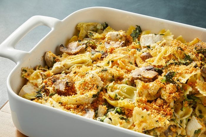 A white casserole dish filled with baked pasta, mushrooms, artichoke hearts, and leafy greens, topped with a crispy breadcrumb crust. The dish is set on a wooden surface.
