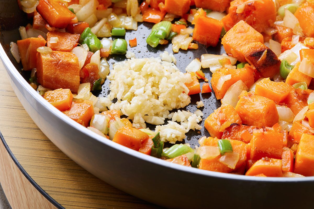A pan filled with diced sweet potatoes, chopped onions, and sliced green beans. A mound of minced garlic is at the center, ready to be mixed. The ingredients are lightly sautéed on a stovetop.